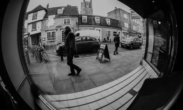 Calle con edificios, coches y gente. Un hombre camina frente a un cartel de "Caffè Nero" con efecto ojo de pez.
