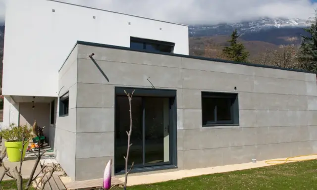 Extension moderne en parpaing gris, 20m2, avec grandes baies vitrées et vue sur montagnes.