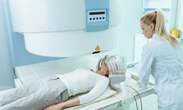 Pacjentka leży w aparacie do rezonansu głowy, a pielęgniarka nadzoruje badanie.