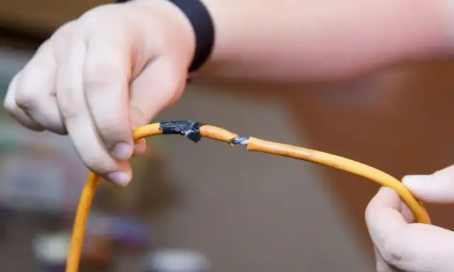 Ręce trzymające pomarańczowy kabel, który jest uszkodzony. Pokazuje, jak połączyć kable bez lutowania, używając taśmy izolacyjnej.
