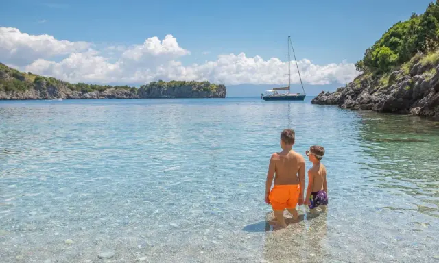 Due bambini esplorano le acque cristalline del Cilento, tra le sue spiagge perfette per famiglie. Una barca a vela all'orizzonte completa il quadro idilliaco.