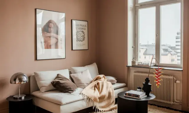 Un divano bianco accogliente con cuscini e una coperta color **rosa pastello** su un tappeto soffice. Alle pareti, due quadri e una finestra con vista.