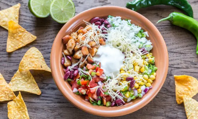 Bunter Taco Salat mit Hähnchen, Reis, Bohnen, Mais, Tomaten, Käse und Sauerrahm. Tortilla-Chips und Limetten daneben.