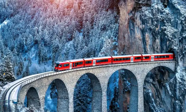 Un treno rosso attraversa un viadotto innevato, parte del suggestivo treno Bernina percorso, tra alberi coperti di neve e rocce imponenti.