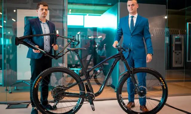 Dwóch mężczyzn prezentuje nowy rower full-suspension. Jeden trzyma ramę, drugi stoi przy gotowym rowerze. To duma firmy Romet.