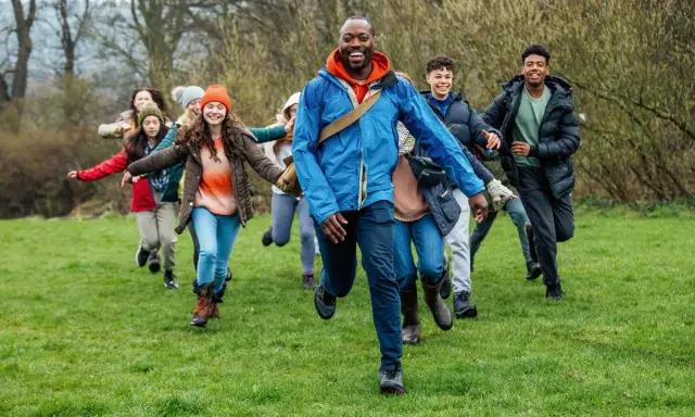 Lehrer und Schüler rennen lachend über eine grüne Wiese. Perfekt, um eine Klassenfahrt zu organisieren und Abenteuer zu erleben!
