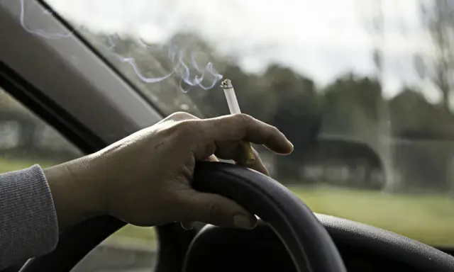 Mano sujetando un cigarrillo encendido sobre el volante. El humo se eleva mientras se puede fumar conduciendo.