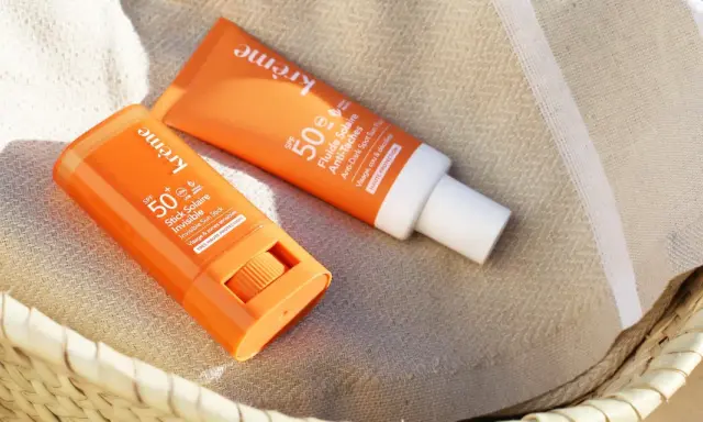 Deux produits solaires, un stick et un fluide, sont posés sur une serviette rayée. Le bouton du stick est visible, prêt à protéger du soleil.
