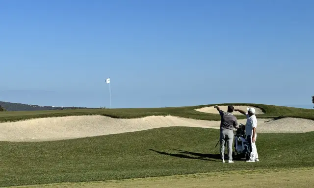 Zwei Golfer planen ihren nächsten Schlag auf dem Valderrama Golfplatz, mit einem Bunker und dem Fahnenmast im Hintergrund.