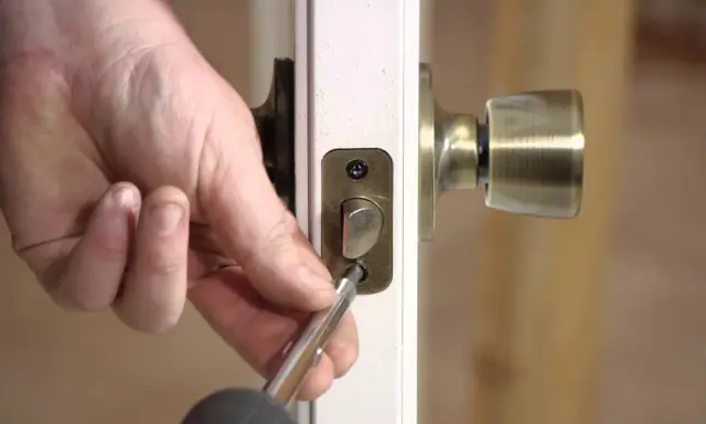 Mano usando un destornillador para desmontar una cerradura de puerta interior, quitando los tornillos de la placa.