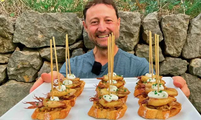 Hombre sonriente presenta cazuelitas de aperitivo con calamares y alioli sobre pan tostado, decoradas con brochetas.