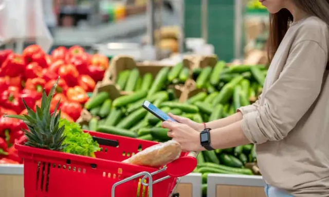 Donna al supermercato con carrello rosso, usa il telefono per i quantificatori spiegazione. Verdure sullo sfondo.