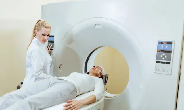 Kobieta w białej bluzie leży na łóżku rezonansu magnetycznego. Technik w białym fartuchu obsługuje panel sterowania.