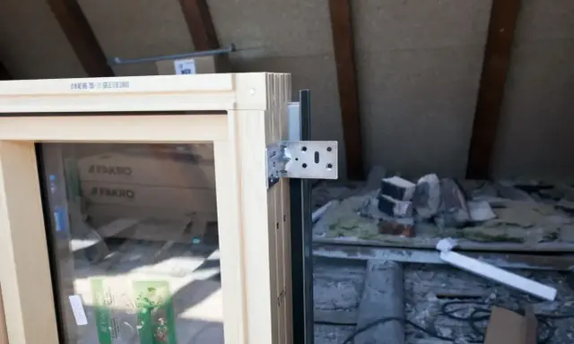 Drewniane okno z metalowym kątownikiem, gotowe do obróbki po montażu. W tle widać materiały budowlane i drewniane belki poddasza.