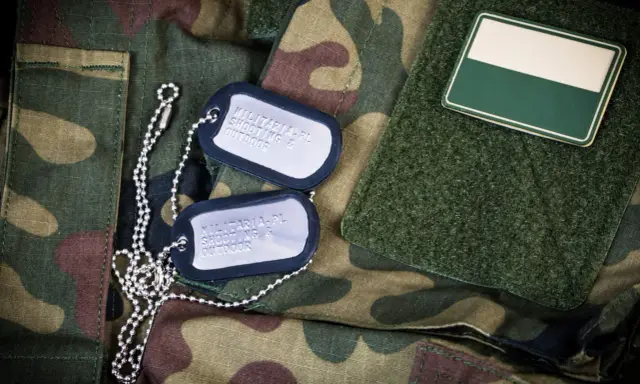 Dwa nieśmiertelniki na mundurze moro. Jeden z nich zawiera napis "MILITARIA.PL SHOOTING & OUTDOOR".