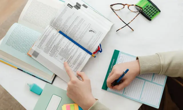 Uczeń pochyla się nad książkami, przygotowując się do egzaminu licencjackiego lub magisterskiego. Na stole leżą notatki, okulary i kalkulator.