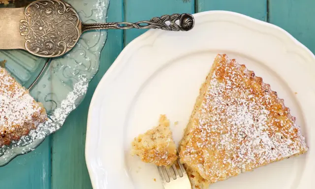 Saftige spanische Mandeltorte, bestäubt mit Puderzucker, auf einem weißen Teller. Ein Stück wurde bereits mit einer Gabel entnommen.