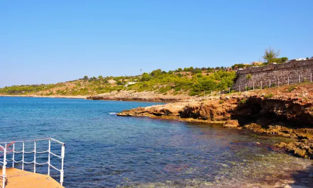 Scogliera rocciosa e mare cristallino a San Gregorio al mare, con vegetazione mediterranea e un muretto in pietra.