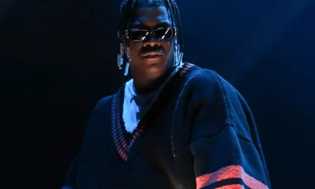 Lil Yachty w okularach i długich kolczykach, w swetrze z pomarańczowymi pasami. Let's start here.