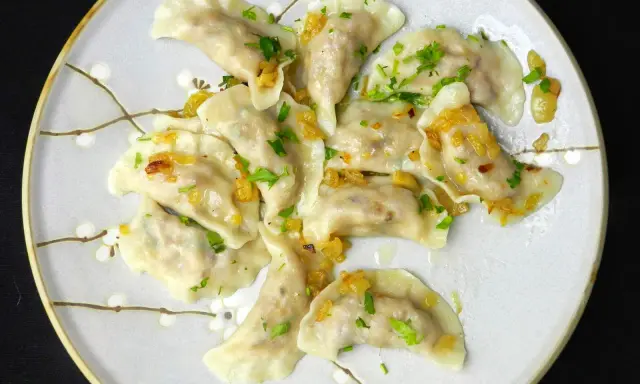 Pyszne pierogi z kaszą gryczaną, okraszone skwarkami i posypane natką pietruszki, na talerzu z delikatnym wzorem kwiatowym.