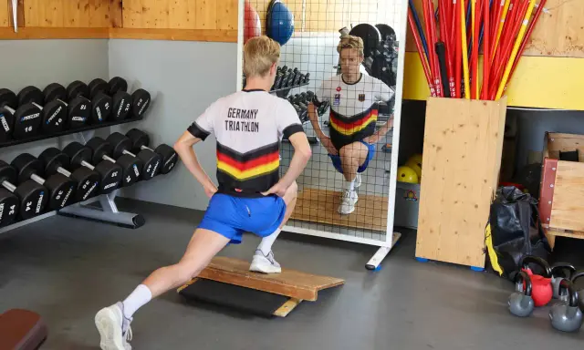 Ein Athlet im "Germany Triathlon"-Shirt macht Ausfallschritte im Athletik Training. Hanteln und Trainingsgeräte im Hintergrund.