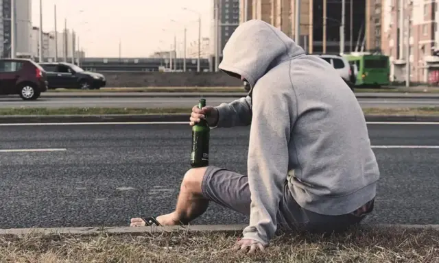 Mężczyzna w kapturze siedzi przy drodze z butelką piwa. Jego postawa sugeruje walkę z nałogiem, być może potrzebuje pomocy terapeuty od uzależnień.