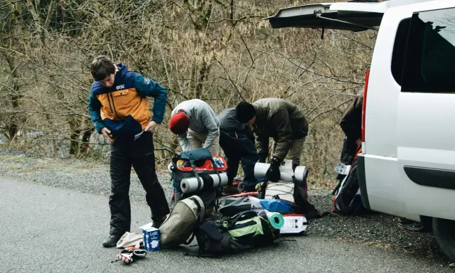 Grupa młodych ludzi przygotowuje sprzęt do wyprawy w góry. Wszyscy szykują się na atrakcje w Tatrach, pakując plecaki i karimaty przy otwartym bagażniku białego vana.