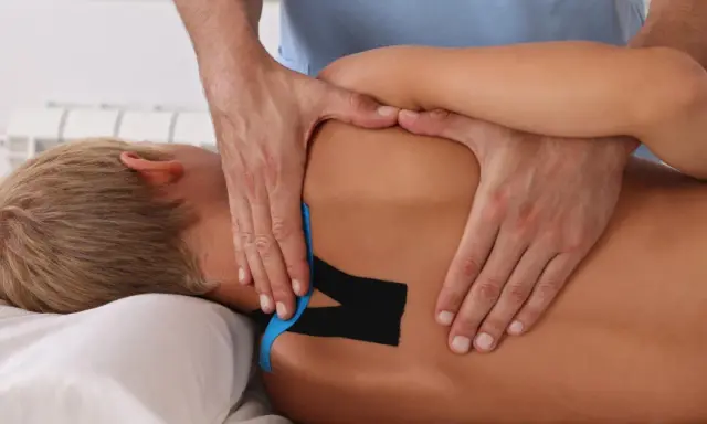 Fizjoterapeuta aplikuje kinesiotaping na plecach pacjenta z chorobą Scheuermanna. Czego nie wolno robić przy tej chorobie? Unikać przeciążeń.