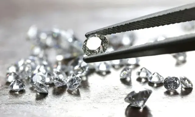 Un diamante etico, scintillante, tenuto da una pinzetta, circondato da altri brillanti su una superficie metallica.