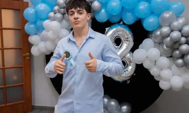 Chłopak w błękitnej koszuli z rozetką "18th Birthday" i balonami.