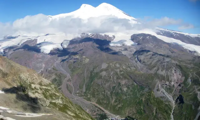 Majestatyczny Elbrus, najwyższy szczyt Europy, góruje nad skalistymi zboczami i lodowcami. W dole widać niewielką osadę, która pokazuje, gdzie leży Elbrus.