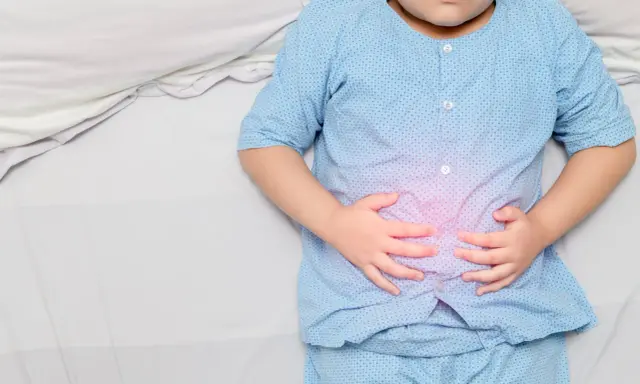 Niño con pijama azul, acostado en cama blanca, se agarra la barriga con las manos. Se visualiza un área rosada en su abdomen, indicando dolor barriga niños.