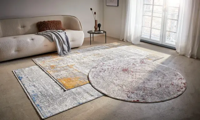 Mattor inspiration: En rund och två rektangulära mattor med abstrakt mönster i grått, vitt, gult och rött ligger på ett betonggolv framför en soffa.
