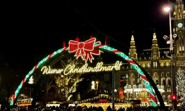 Wiedeński jarmark świąteczny w blasku świateł. Tłumy ludzi spacerują pod łukiem z napisem "Wiener Christkindlmarkt", w tle widać zabytkowe budynki i choinkę.