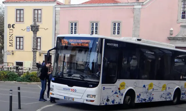 Ein Scott-Bus in Portugal hält an einer Haltestelle. Touristen steigen ein, während im Hintergrund bunte Gebäude und ein historischer Brunnen zu sehen sind.