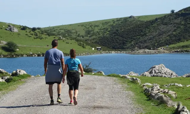 Ein Paar wandert auf einem Schotterweg am Ufer eines Sees. Grüne Hügel und Felsen prägen die **Spanien Natur**.