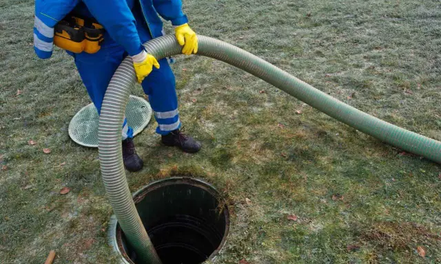 Pracownik w niebieskim kombinezonie i żółtych rękawicach wykonuje czyszczenie szamba, wkładając gruby wąż do otwartego włazu.