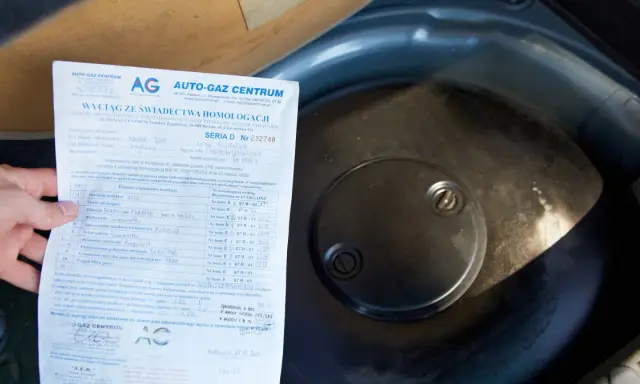 Fragment dokumentu z danymi homologacji butli LPG obok okrągłej pokrywy w bagażniku samochodu.
