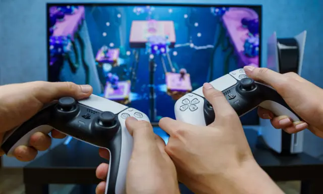 Dwie osoby grają w grę na konsoli PlayStation 5, trzymając kontrolery DualSense.