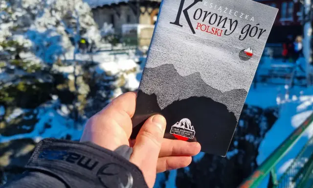 Dłoń trzyma książeczkę "Korona Gór Polski" na tle zimowego krajobrazu.