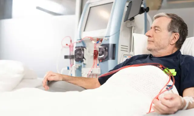 Starszy mężczyzna leży pod kocem, podłączony do aparatury do dializy. Jego ręka spoczywa na podłokietniku, a obok widać maszyny i czerwone wężyki.