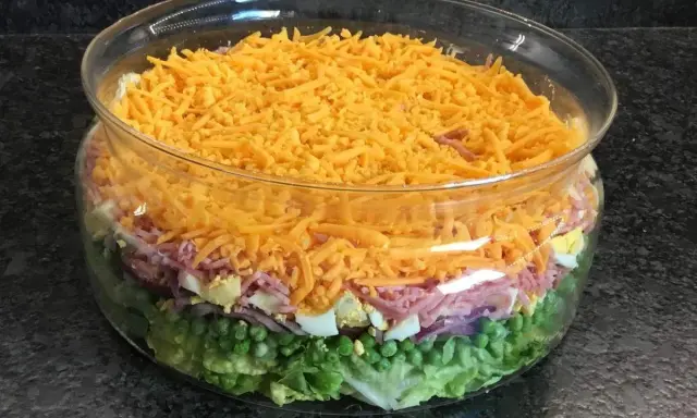 Une salade 7 étages colorée dans un grand bol en verre, avec des couches de laitue, petits pois, oignons rouges, jambon, œufs durs et fromage râpé.