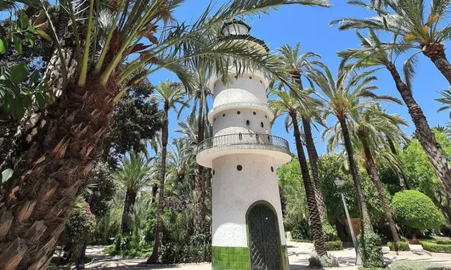 Torre blanca con balcón y puerta verde, rodeada de palmeras y vegetación exuberante. Un lugar encantador que ver en Elche.