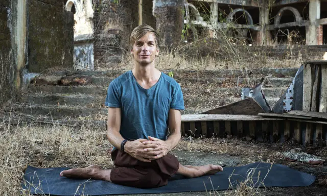 Man i blå tröja sitter i lotusställning på en yogamatta i ruiner. En stunds natthiko meditation i en övergiven miljö.