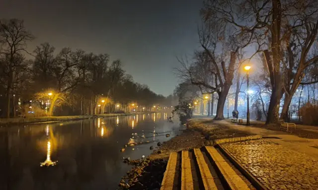 Mglisty wieczór nad **Bydgoszczą**, latarnie oświetlają spokojną **rzekę** i aleję drzew.