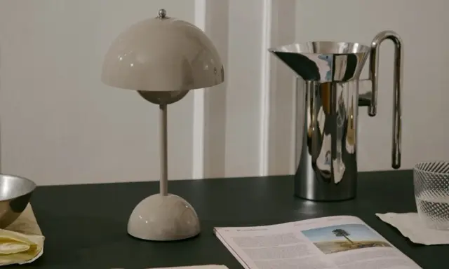 En beige lampa i studio lawahl-stil står bredvid en blank vattenkanna. En tidning med ett träd på omslaget ligger framför.