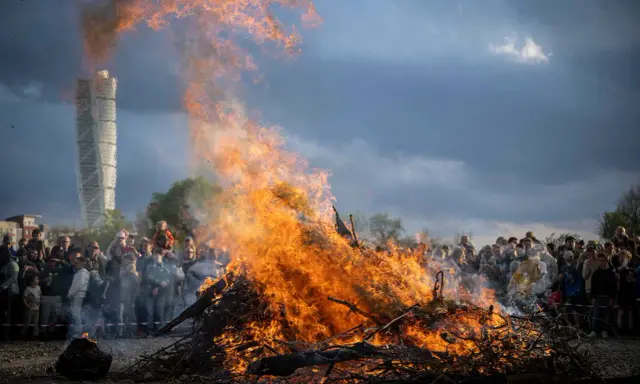 Stort bål med flammor som slår upp mot en dramatisk himmel. En folkmassa samlas för att bevittna ett traditionellt valborg firande.