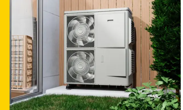 Unidad exterior de una bomba de calor para calefacción, instalada junto a una pared de madera y rodeada de vegetación.