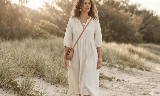 A woman walks on a beach in a flowing white dress, a tan crossbody bag, and gold jewelry. Her look embodies a natural, soft style, perfect for a David Kibbe test.