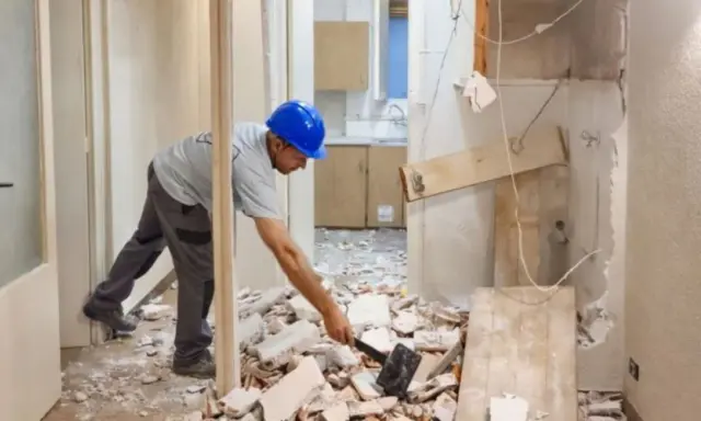 Obrero con casco azul trabajando en la demolición de un tabique, rodeado de escombros.